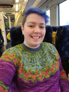 A selfie of a short & blue haired person wear a colourful sunset highway sweater